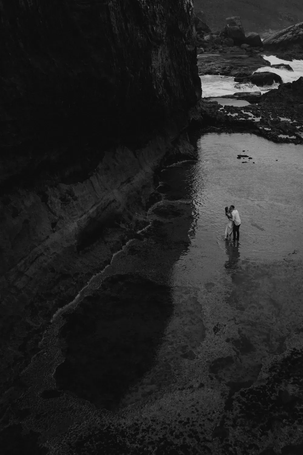 Black and white photograph of a couple embracing in a rock pool below coastal cliffs