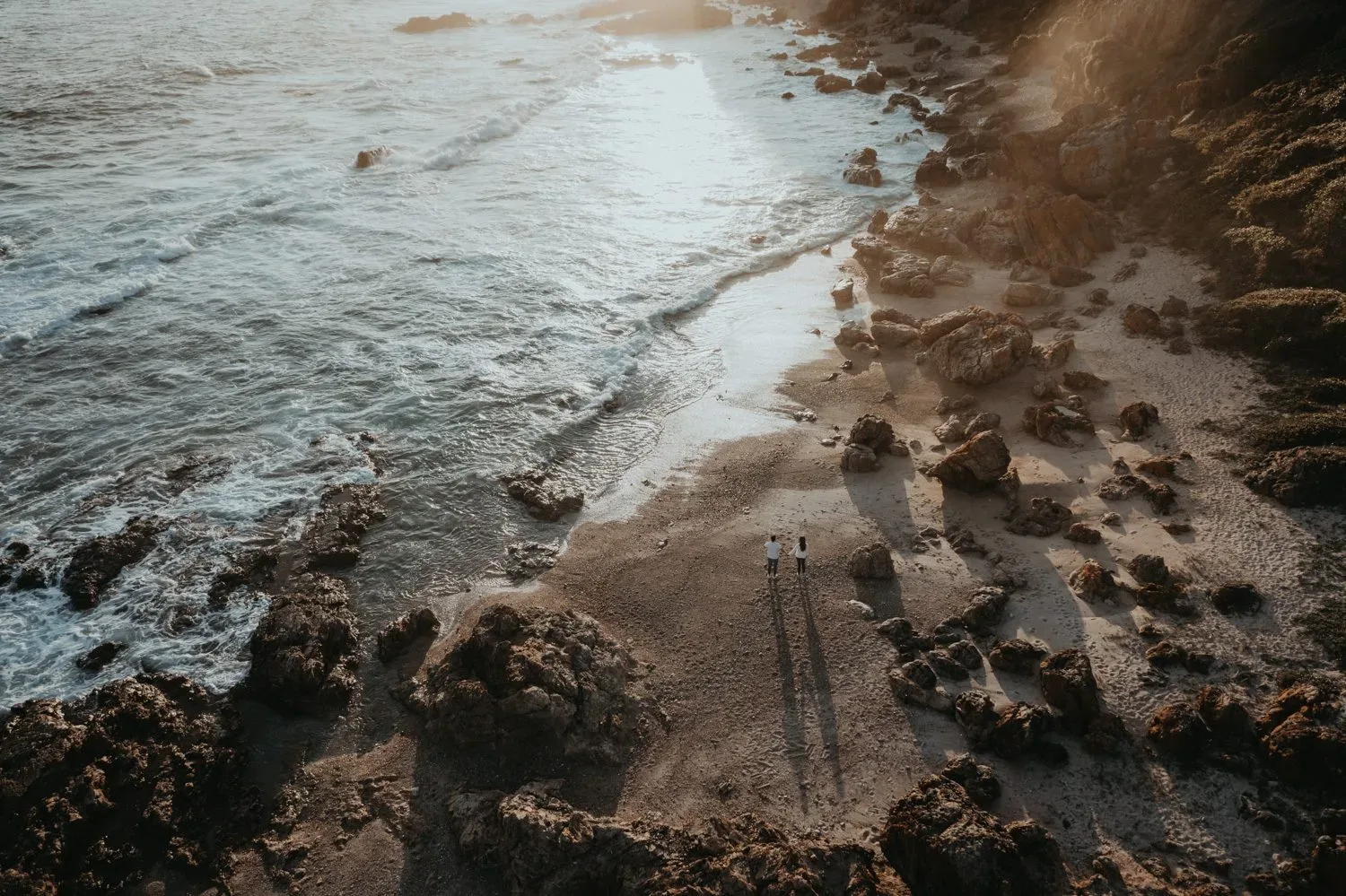 A couple walks along a rocky beach seen from above in warm evening light