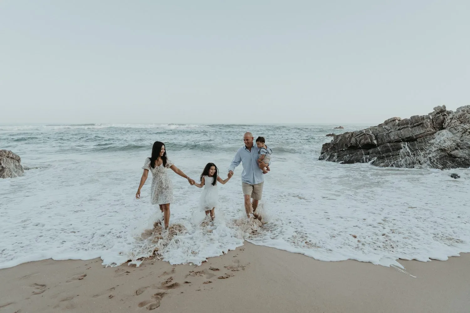 A family holds hands in the shallow surf beside coastal rocks