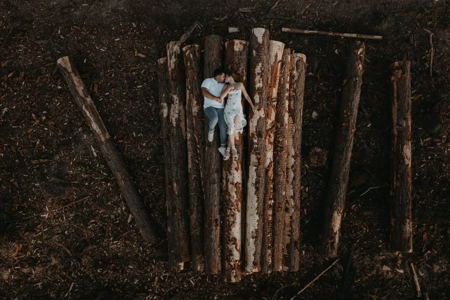 A couple lies together on stacked timber in a forest clearing